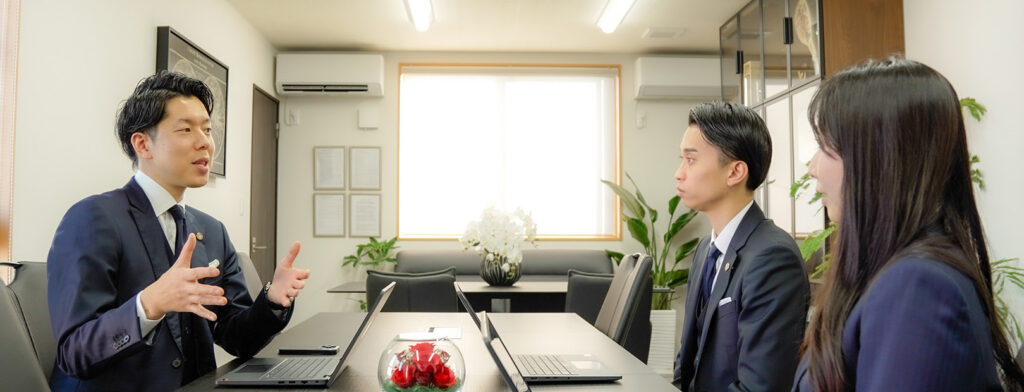 会議室でのチームミーティング。代表の榊氏がスタッフ二人に語りかけ、互いに真剣な表情で意見を交わしている。