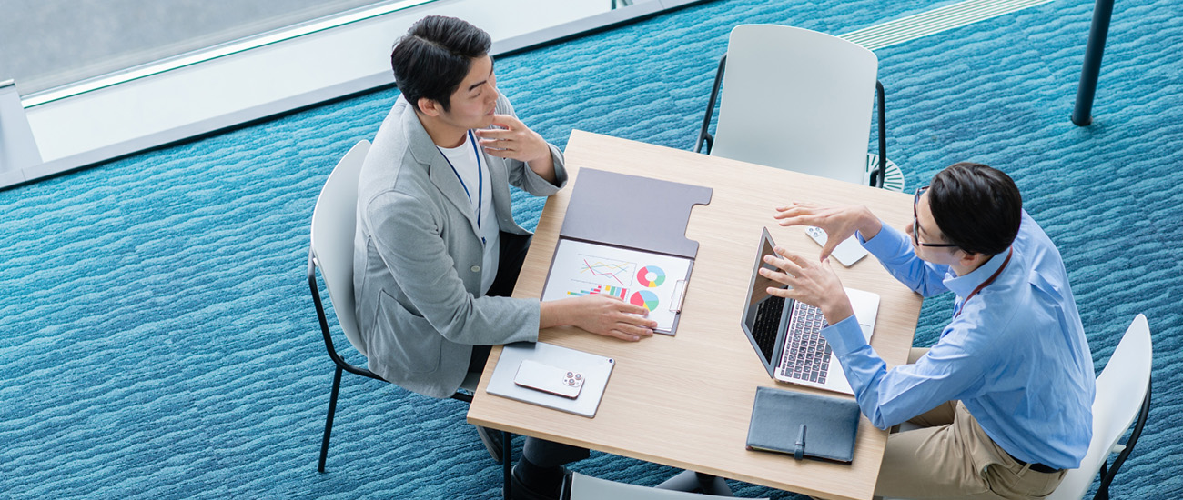 テーブルに資料やタブレットを広げ、会話している二人の男性