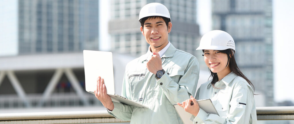 建設現場を背景に、ヘルメットを着用して笑顔を見せる男女の作業員