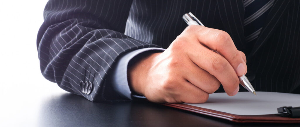 スーツ姿の人物が万年筆で書類に署名をする手元のアップ